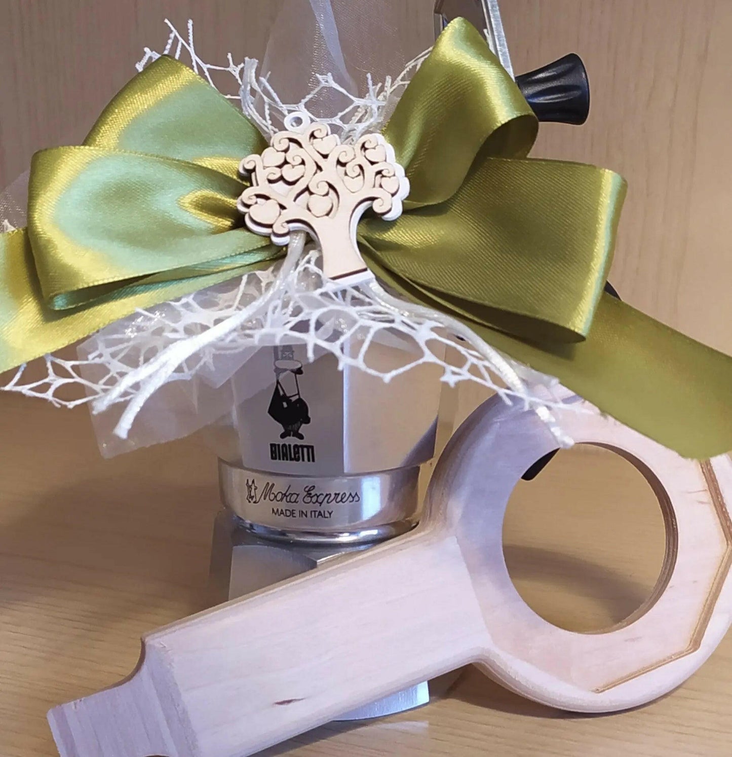 Aprimoka come idea regalo con Caffettiera Bialetti Moka Express 3 tazze sopra la caffettiera vi è posta una bomboniera con fiocco verde oliva ed al centro un alberello inciso nel legn. L'aprimoka si trova alla base della caffettiera e vi è appoggiato.